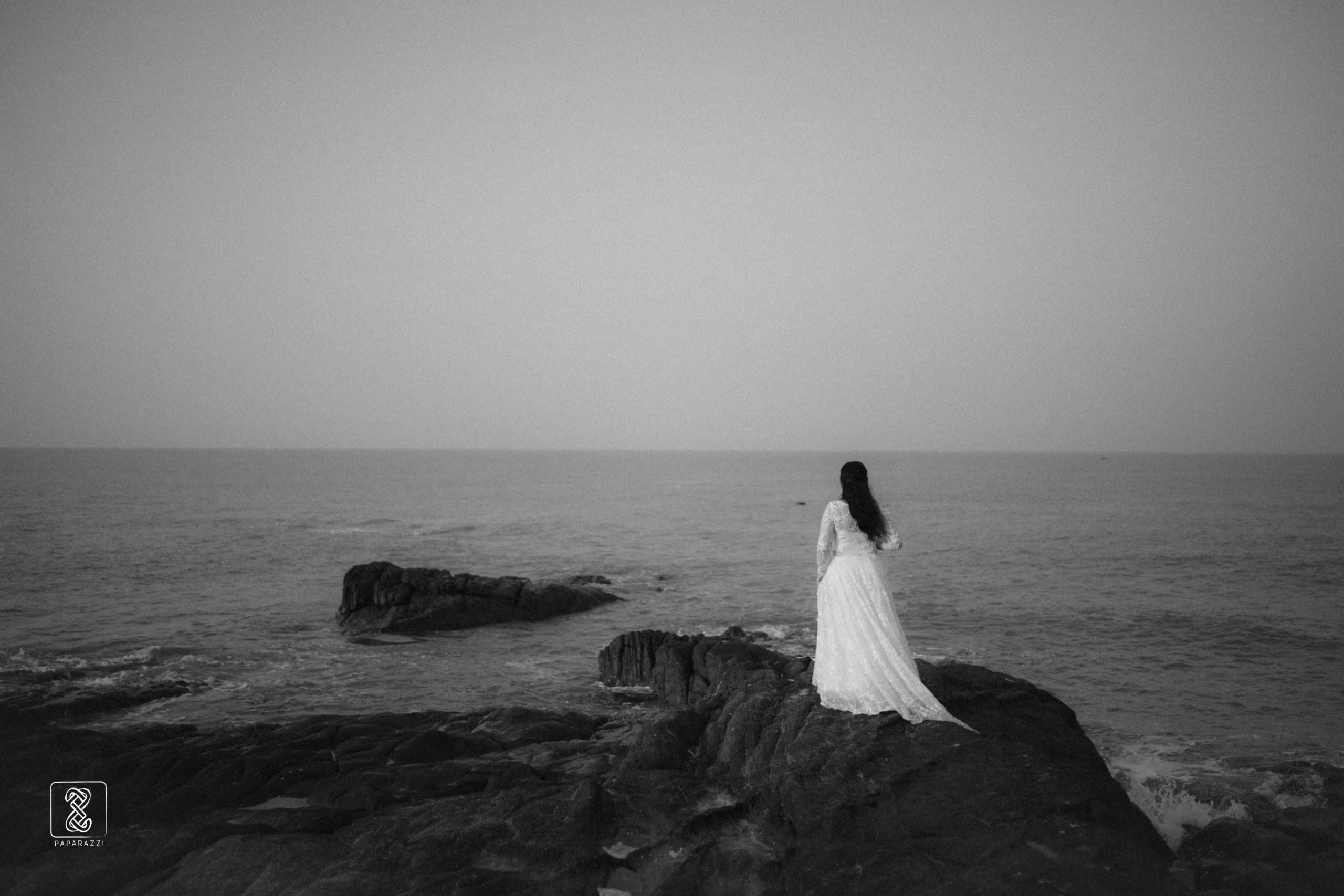 Bride in a white lace dress standing on coastal rocks overlooking the ocean.